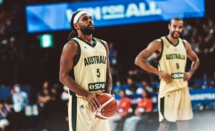 An Indigenous basketball player wearing an Australian uniform holds a basketball and prepares to shoot. There is a teammate and a stadium crowd in the background.